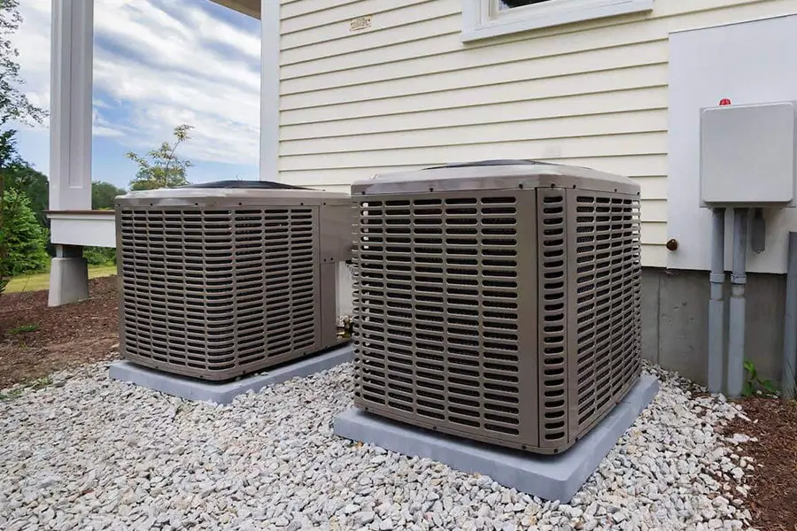 Two outdoor air conditioning condenser units installed on pads over gravel beside a house.
